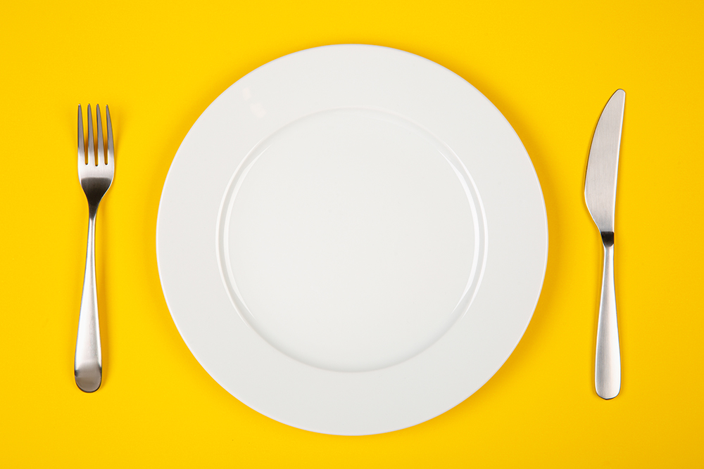 Silver fork and knife on either side of white plate, on a yellow background